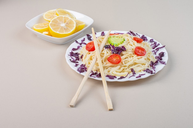 A plate of cooked noodles with vegetables and fresh sliced lemon.