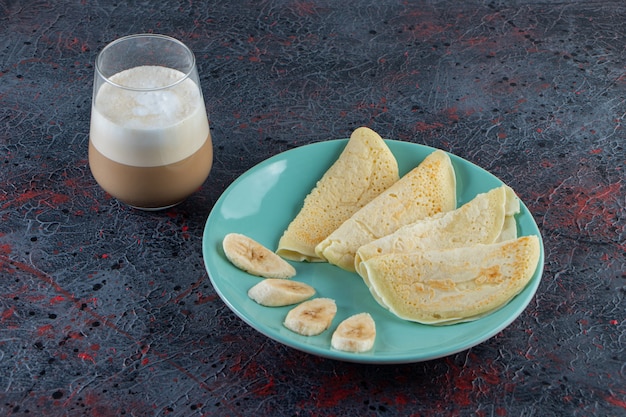 Plate of crepes and sliced bananas with glass of milk coffee on dark surface. 