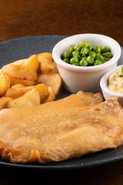 Plate of fish and chips with sliced fried fish and pub table closeup with tartar sauce and green peas closeup portrait on breading