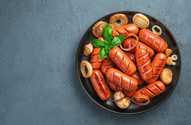 A plate of fried sausages with onions, garlic and basil on a dark gray background. Top view, space for copying.