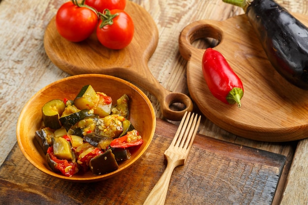 A plate of grilled vegetables next to tomatoes and eggplant and peppers on wooden boards next to a wooden fork