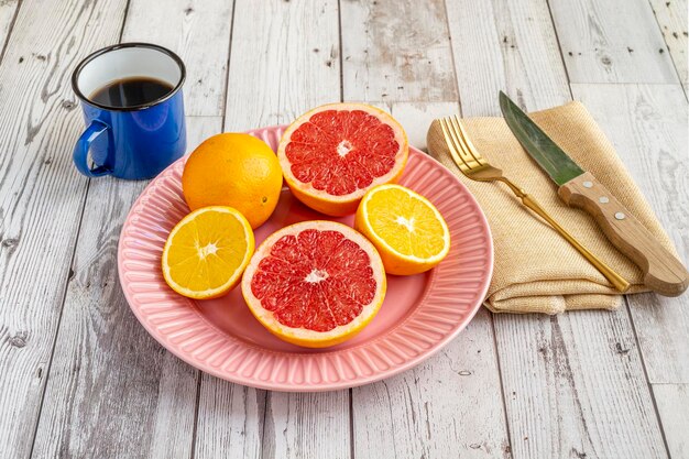 Plate of halved citrus fruits oranges and grapefruits with coffee and cutlery on light wooden table