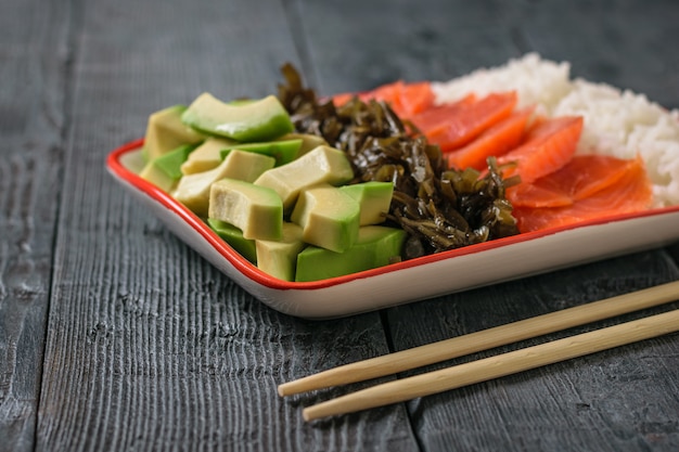 A plate of Hawaiian rice, avocado, salmon and kelp on a black rustic table. The view from the top.