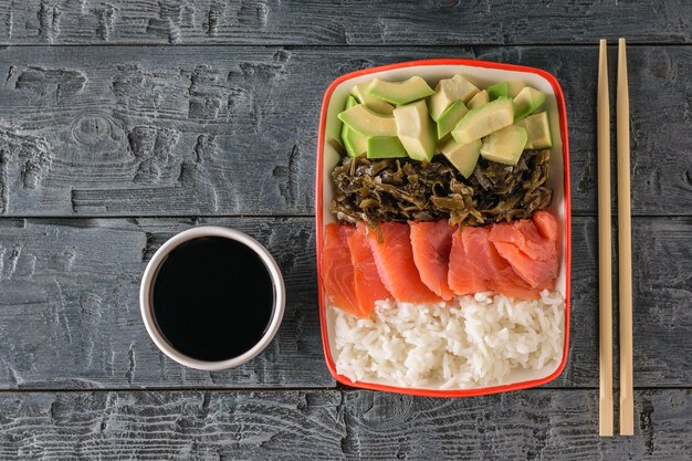 A plate of Hawaiian rice, avocado, salmon and kelp and soy sauce on a black dark table. The view from the top.