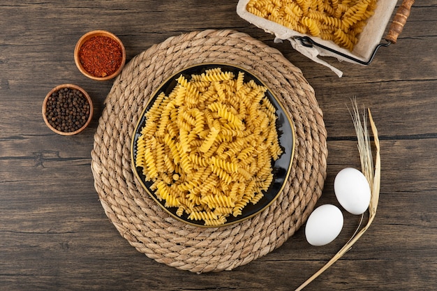 Plate of raw fusilli, condiments and raw eggs on wooden surface.