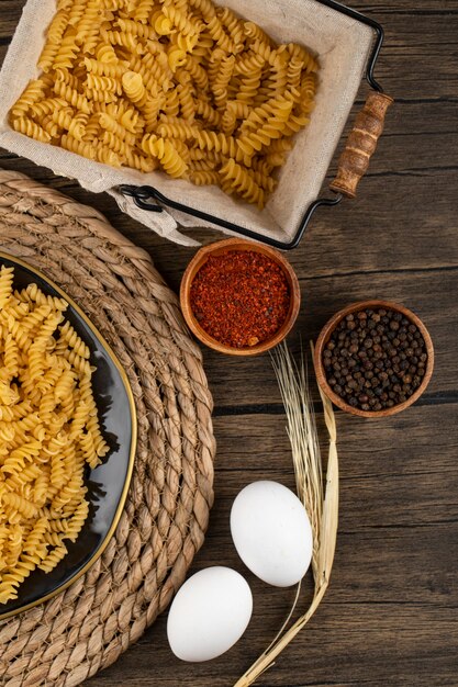 Plate of raw fusilli, condiments and raw eggs on wooden surface. 