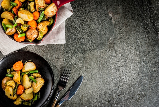 Piatto e padella con verdure autunnali fritte di stagione (zucchine, patate, carote, fagioli), sul tavolo di pietra nera