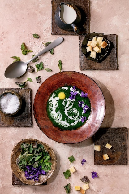 Тарелка супа-пюре из весенней травяной крапивы, подается с перепелиным желтком, фиалками, цветами, кремовыми гренками и молодыми листьями крапивы на коричневой керамической плитке. Розовая текстура фона. Здоровая пища.