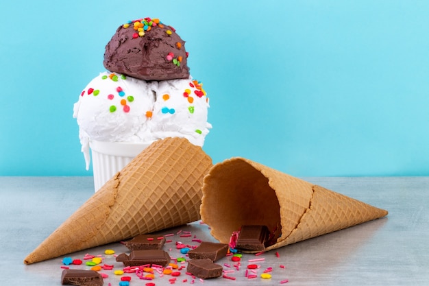 Plate of vanilla and chocolate ice cream scoops swith sprinkles chocolate pieces and waffle cones on a blue.