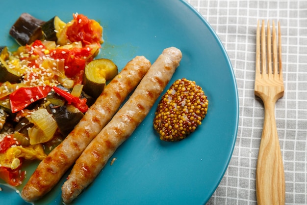 A plate of vegetables tomatoes zucchini eggplant with grilled sausages next to a wooden fork closeup