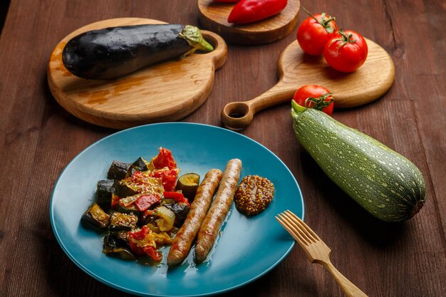 A plate of vegetables with grilled sausages on the table next to tomatoes zucchini eggplant on wooden boards and a wooden fork