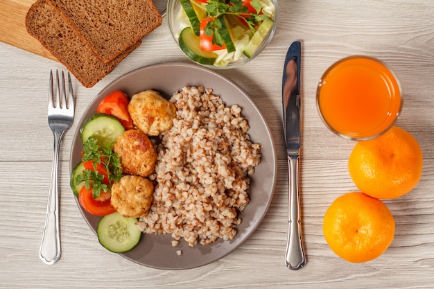 Тарелка с гречкой, котлеты из жареного мяса, кусочки свежих огурцов, помидоры и свежая петрушка, нож и вилка, хлеб, стакан апельсинового сока и апельсины. Вид сверху.