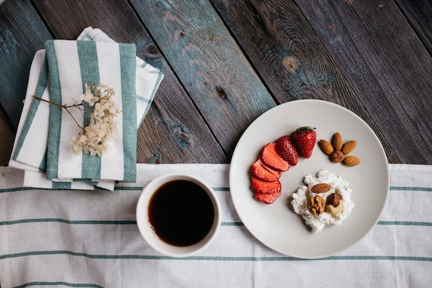 Plate with cottage cheese, strawberries and nuts, a cup of coffee and towels on wooden table, healthy food, breakfast