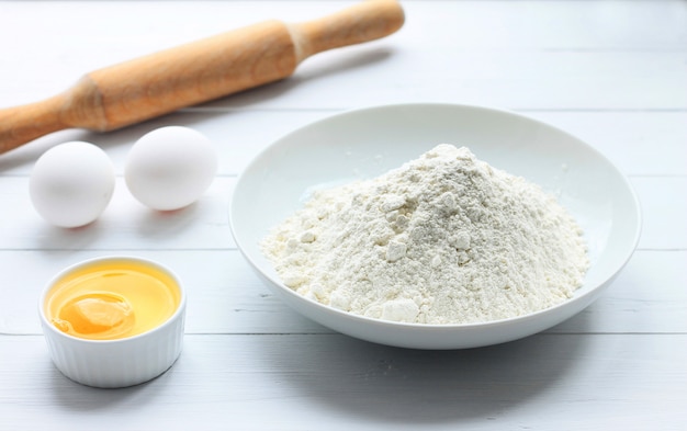 A plate with flour, eggs, a rolling pin on a white wooden background. 