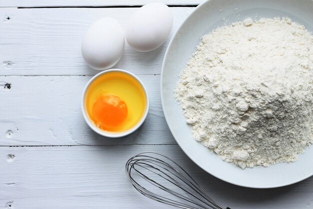 A plate with flour, eggs, a whisk on a white wooden background. 