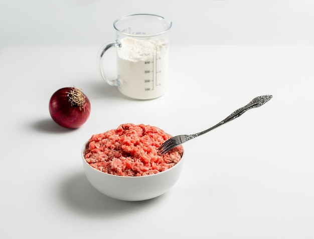 신선한 갈은 쇠고기와 흰색 배경에 밀가루를 넣은 붉은 양파 측정 컵이 있는 접시