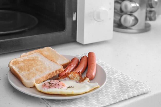 Plate with fried egg sausages and toasts near microwave on table