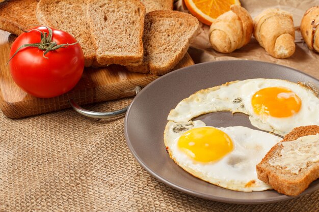 Plate with fried eggs, toast with butter, croissants, bread and fresh tomato on wooden cutting board on table covered with sackcloth