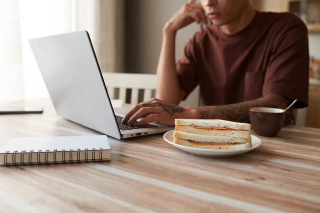 自宅でラップトップに取り組んでいる学生の机の上にハムとチーズのサンドイッチでプレート