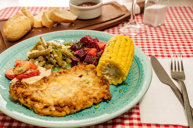 Тарелка с мясным шницелем, томатным салатом, зеленой фасолью, свеклой, яйцом вкрутую и кукурузными початками на традиционном столе в трактире или ресторане. Горизонтальный вид