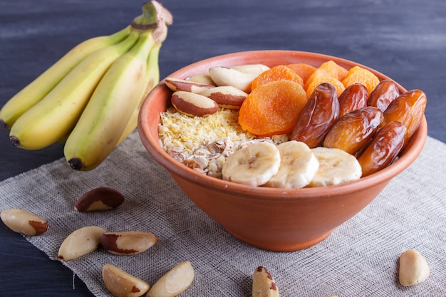 Un piatto con muesli, banana, albicocche secche, date, noci del brasile su un fondo di legno nero.