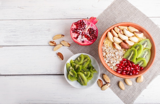 A plate with muesli, kiwi, pomegranate, Brazil nuts on a white wooden background.