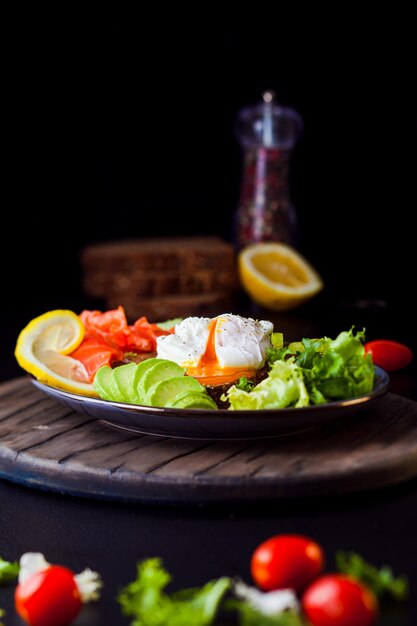 Plate with pashot eggs salmon avocado and vegetables delicious\
breakfast on dark wooden board and black background variety in\
eating for good mood