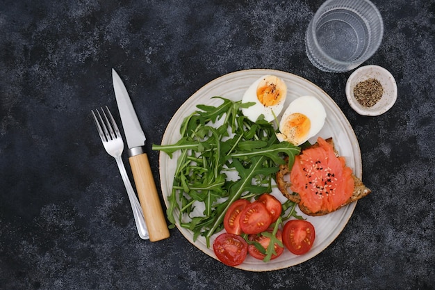 A plate with a salmon sandwiche arugula tomatoes boiled egg with glass of water on black background