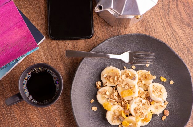 Plate with sliced banana, granola, candied fruit and honey, toast and a cup of coffee.