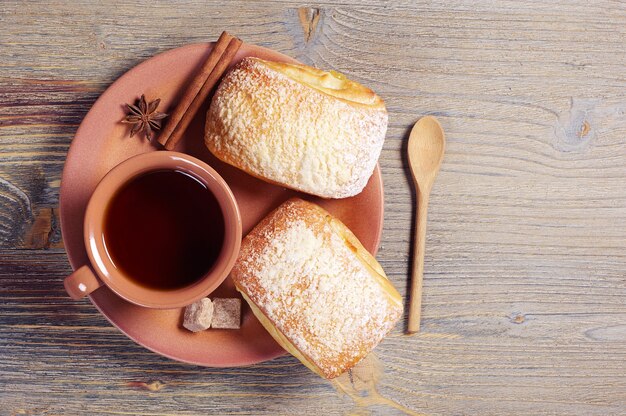 Plate with two fresh buns and cup of hot tea on rustic wooden table, top view