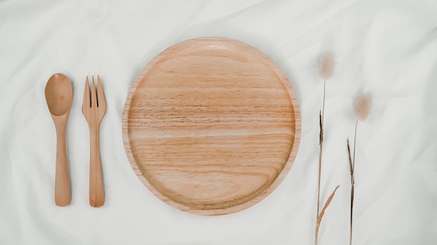 Тарелка деревянная, ложка деревянная и вилка деревянная с сухим цветком Хвост Кролика на белой ткани. Вид сверху сервировки стола на белом фоне