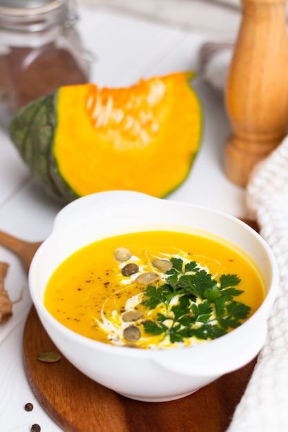 A plate of yellow pumpkin cream soup with cream herbs and seeds on a white table near ginger raw pumpkin and a wooden spoon Vertical