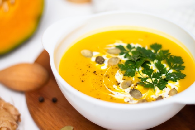 A plate of yellow pumpkin cream soup with cream herbs and seeds on a white table near ginger raw pumpkin and a wooden spoon