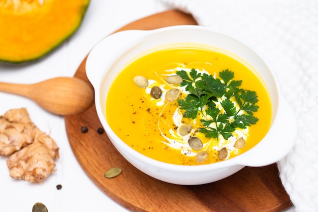 A plate of yellow pumpkin cream soup with cream herbs and seeds on a white table near ginger raw pumpkin and a wooden spoon