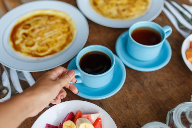 Plates with banana pancakes, tropical fruits and two cups of coffee on wooden table.