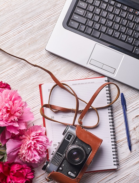 Platliggend damesbureau. Vrouwelijke werkruimte met laptop, roze pioenrozen boeket, camera en koffie op witte achtergrond. Bovenaanzicht vrouwelijke achtergrond.