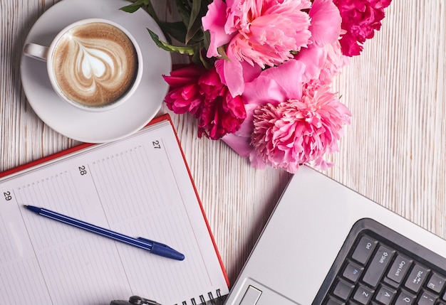 Platliggend damesbureau. Vrouwelijke werkruimte met laptop, roze pioenrozen boeket en koffie op witte achtergrond. Bovenaanzicht vrouwelijke achtergrond.