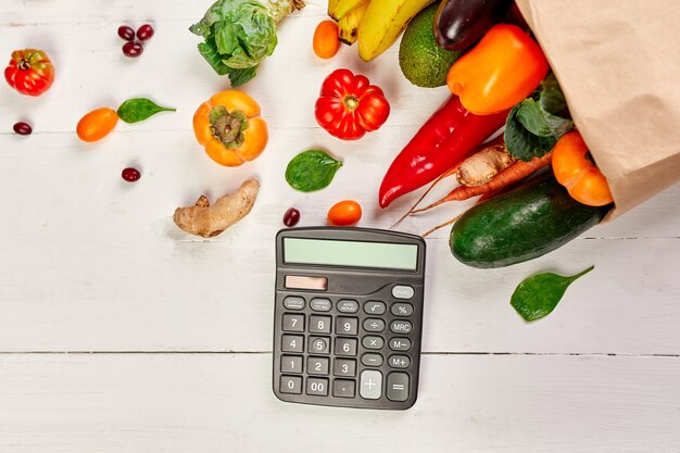 Foto platte papieren boodschappentas met assortiment van verse groenten en fruit en rekenmachine, bio-gezond, biologisch voedsel op witte achtergrond, supermarktstijl, kruidenierswinkel, dieet vegetarisch voedsel.