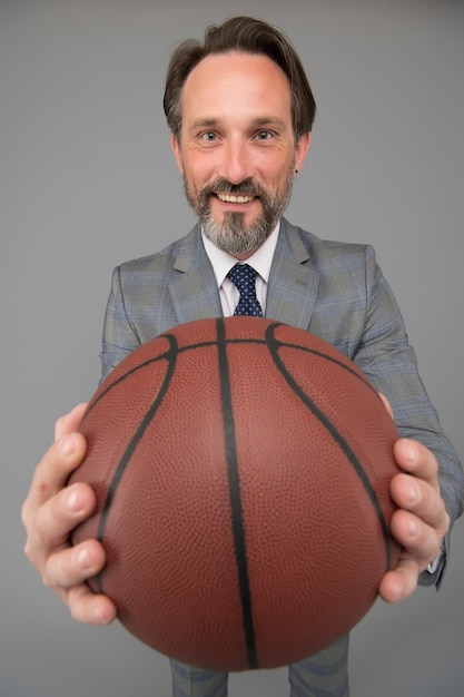 幸せになるバスケットボールをする。幸せなビジネスマンはバスケットボールのボールを保持します。バスケットボールのコーチの灰色の背景。バスケットボールのコーチング。ビジネスとスポーツの競争。競争力のあるゲーム。一生懸命遊べ。