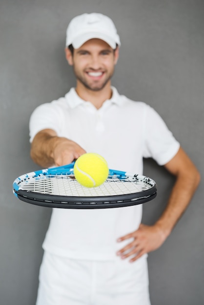 Play with me! Happy young man in sports clothes stretching out tennis racket with ball and smiling while standing against grey background