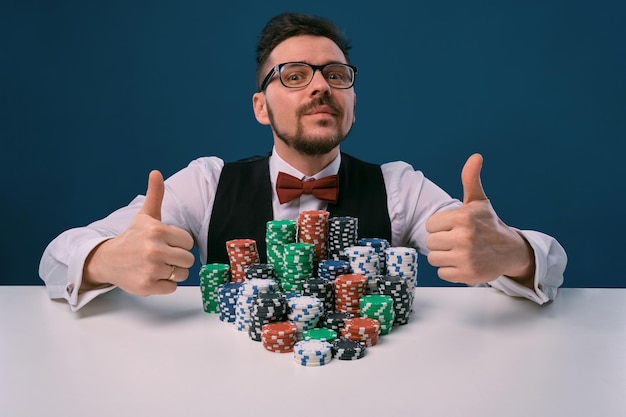 Player in glasses black vest and shirt sitting at white table with stacks of chips on it posing on b...
