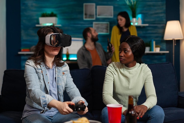 Player woman with virtual reality headset holding gaming joystick playing videogames during online competition. Group of multi-ethnic friends supporting her enjoying free time together