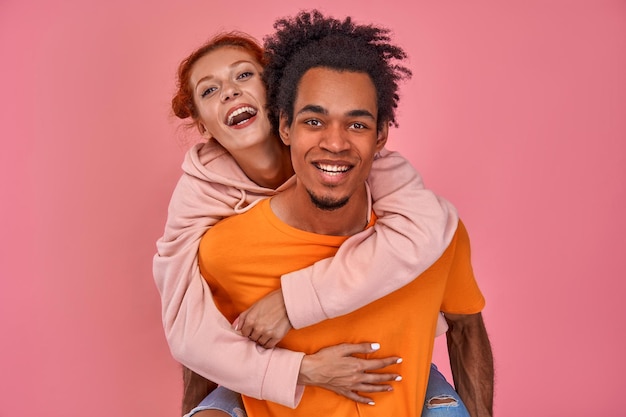 遊び心のあるアフリカ系アメリカ人の男は、ピンク色の上で幸せに再髪のガールフレンドの笑顔にピギーバックに乗る