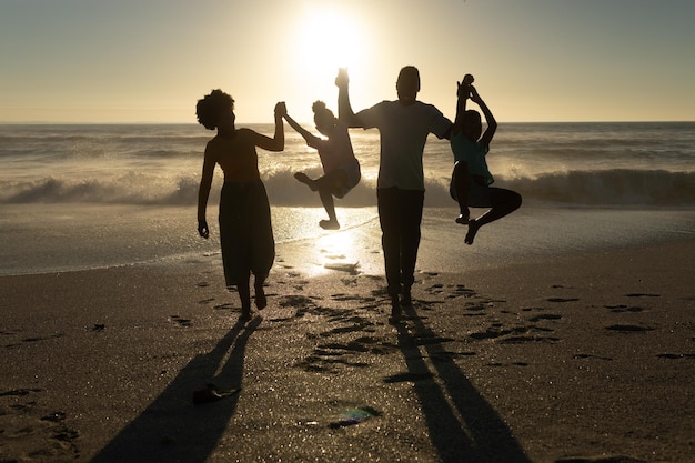 Playful african american parents holding hands of children at beach against sky during sunset