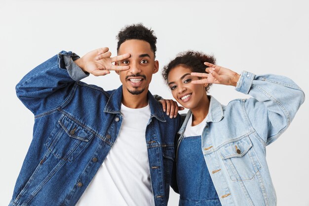 Playful african couple in denim shirts showing peace gestures  over grey wall