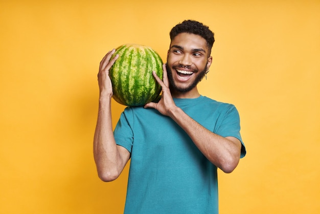 Playful african man carrying watermelon on shoulder while standing against yellow background