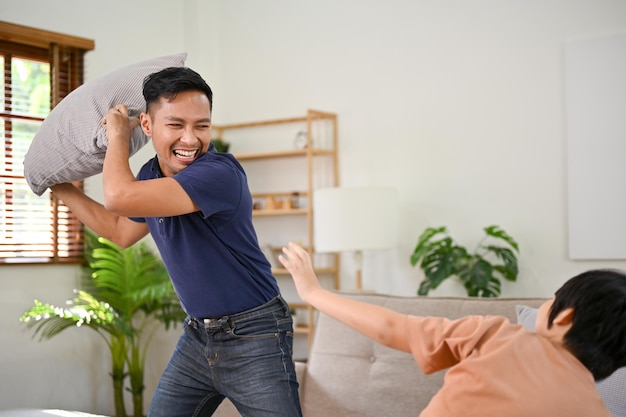 Playful Asian dad enjoying playing pillow fight with his little son in the living room