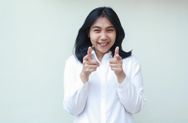 Playful asian young business woman excited pointing forefinger and thumbs up to camera wearing white formal suit shirt its you gesture isolated in white background adevertisement concept