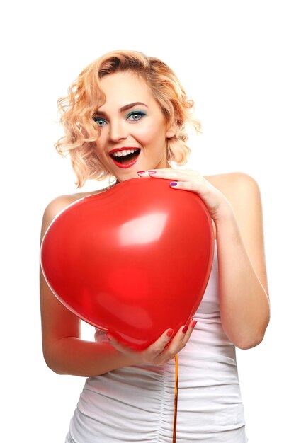 Playful attractive blond girl holding red heart balloon isolated on white