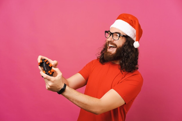 Playful bearded man with Christmas hat is playing with joystick in pink studio